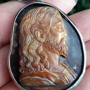 May include: A silver pendant featuring a carved cameo of Jesus Christ's profile. The cameo is made of a brown shell-like material and is set in a silver frame.