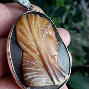 May include: A silver pendant with a cameo of a woman's face carved from shell. The cameo is set in a silver frame and has a dark background.