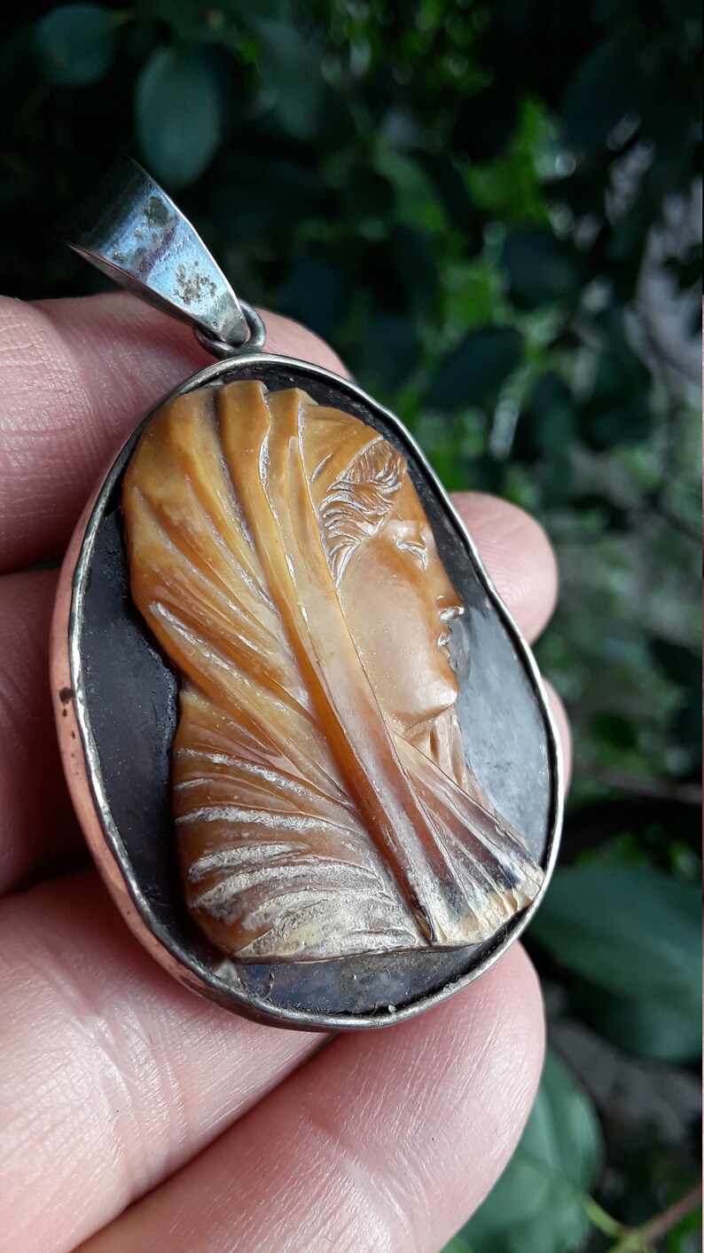 May include: A silver pendant with a cameo of a woman's face carved from shell. The cameo is set in a silver frame and has a dark background.