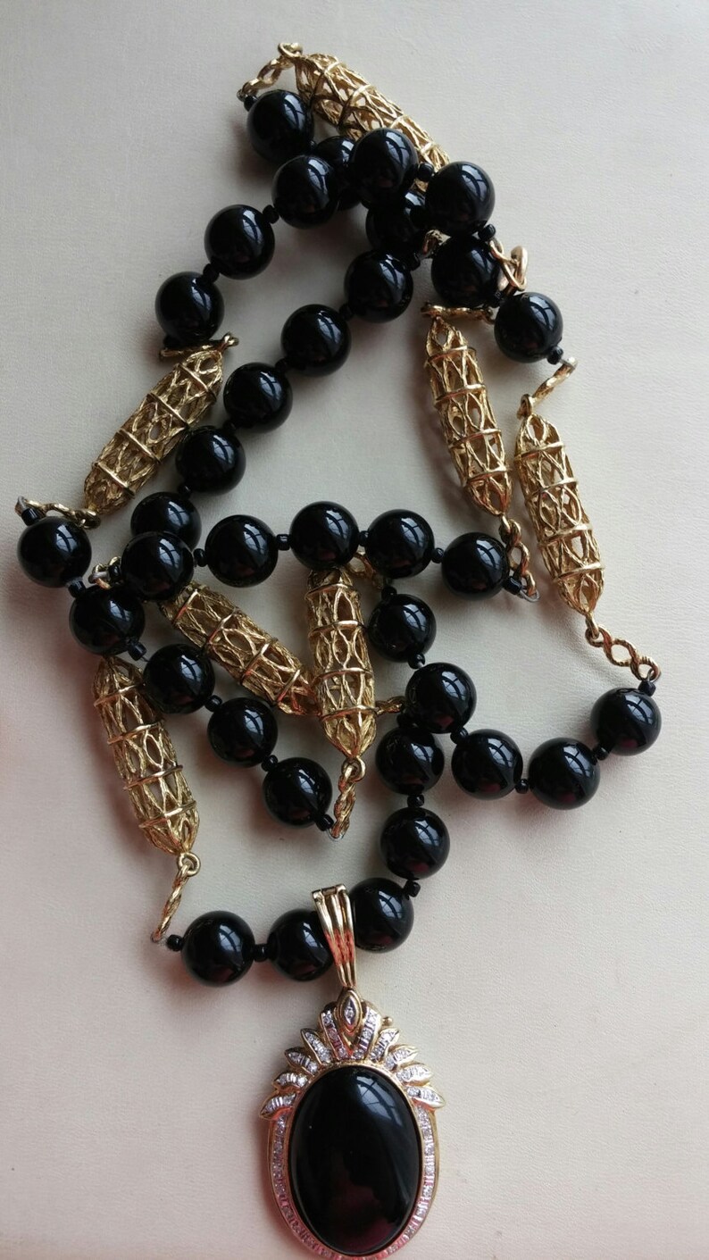 May include: A black and gold necklace with a large black oval pendant. The pendant is set with small clear stones and has a gold filigree design.