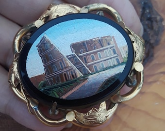 May include: A gold-toned brooch with a black oval cameo featuring a mosaic image of a building. The cameo is surrounded by a decorative gold frame with leaf-like details.