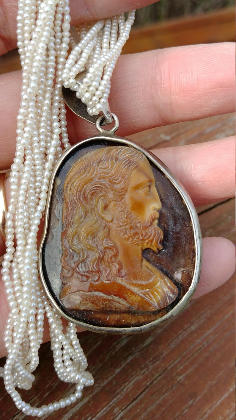 May include: A silver chain necklace with a cameo pendant featuring a carved profile of Jesus Christ. The cameo is set in a silver oval frame and is surrounded by a strand of white pearls.