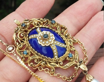 May include: A gold pendant necklace with a blue enamel oval centerpiece surrounded by pearls and blue gemstones. The pendant is attached to a delicate gold chain.