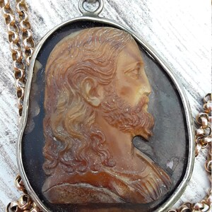 May include: A silver pendant necklace with a cameo of Jesus Christ. The cameo is made of a brown, translucent material and features a detailed profile of Jesus with long hair and a beard. The pendant is attached to a gold chain.