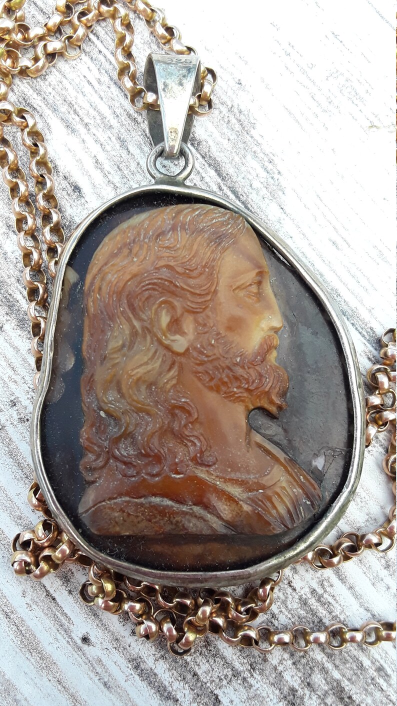 May include: A silver pendant necklace with a cameo of Jesus Christ. The cameo is made of a brown, translucent material and features a detailed profile of Jesus with long hair and a beard. The pendant is attached to a gold chain.