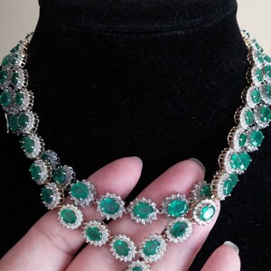 May include: A silver and gold necklace with a pendant featuring a cluster of emerald green gemstones surrounded by smaller diamonds. The necklace is made up of a series of oval-shaped emerald green gemstones surrounded by smaller diamonds.
