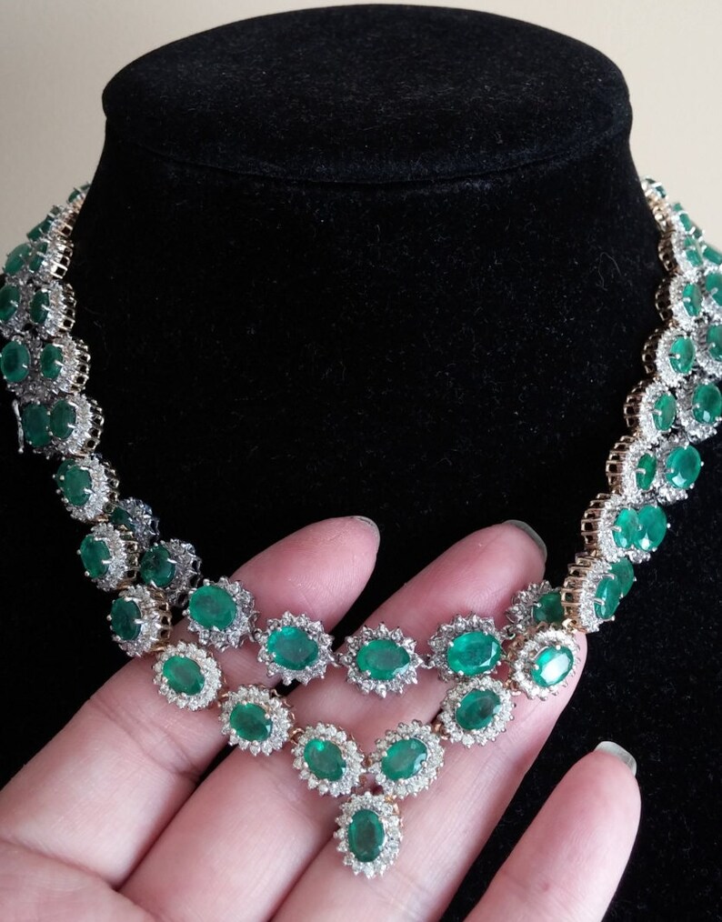 May include: A silver and gold necklace with a pendant featuring a cluster of emerald green gemstones surrounded by smaller diamonds. The necklace is made up of a series of oval-shaped emerald green gemstones surrounded by smaller diamonds.