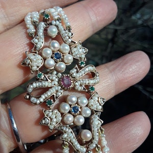 K&ouml;nnte beinhalten: Ein zartes Armband aus Gold und Perlen mit einem floralen Design. Das Zentrum des Designs zeigt eine Ansammlung von Perlen mit einem Rubin- und Saphir-Edelsteinakzent. Das Armband ist mit gr&uuml;nen Edelsteinen und kleinen Perlen verziert.