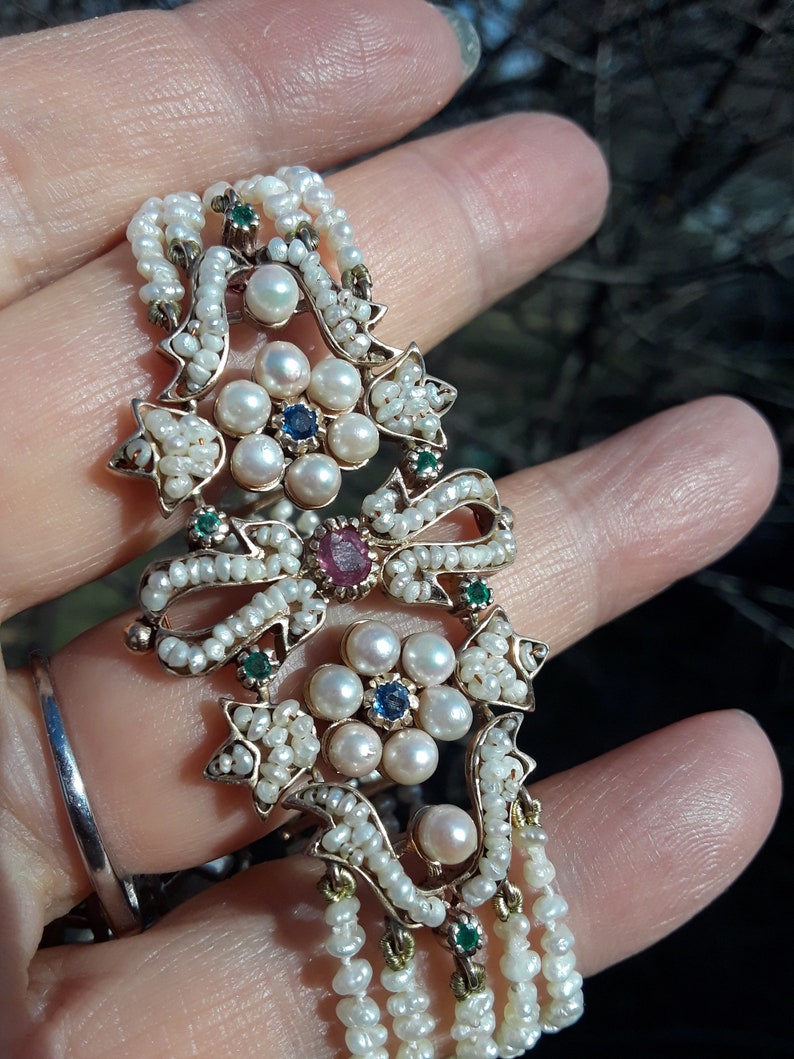 K&ouml;nnte beinhalten: Ein zartes Armband aus Gold und Perlen mit einem floralen Design. Das Zentrum des Designs zeigt eine Ansammlung von Perlen mit einem Rubin- und Saphir-Edelsteinakzent. Das Armband ist mit gr&uuml;nen Edelsteinen und kleinen Perlen verziert.