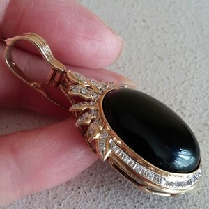 May include: A gold pendant with a black oval gemstone surrounded by a halo of small diamonds. The pendant has a bail for attaching to a chain.