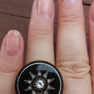 K&ouml;nnte beinhalten: Ein schwarzer Ring mit einem gro&szlig;en, runden, schwarzen Stein, der mit einem Sternendesign aus kleineren Steinen besetzt ist. Der Ring wird an einem Finger mit klarem Nagellack getragen.