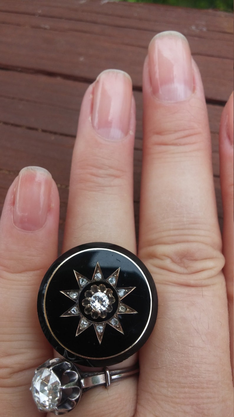 K&ouml;nnte beinhalten: Ein schwarzer Ring mit einem gro&szlig;en, runden, schwarzen Stein, der mit einem Sternendesign aus kleineren Steinen besetzt ist. Der Ring wird an einem Finger mit klarem Nagellack getragen.