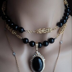May include: A layered necklace with black beads and a gold chain. The pendant features a large oval black stone surrounded by smaller clear stones.