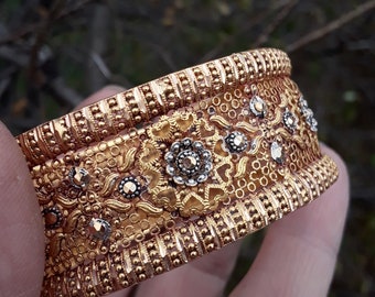 May include: A gold bangle bracelet with intricate floral designs and small, clear stones. The bracelet has a textured, beaded edge.
