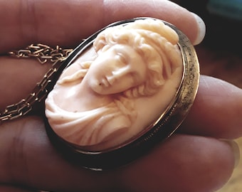 May include: A gold-toned pendant necklace with a cameo featuring a carved portrait of a woman with a detailed hairstyle. The cameo is set in a oval-shaped frame.