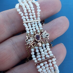 K&ouml;nnte beinhalten: Ein goldenes Armband mit einem roten Edelstein und Perlenakzenten. Das Armband besteht aus mehreren Reihen von Perlen und hat einen dekorativen Verschluss.