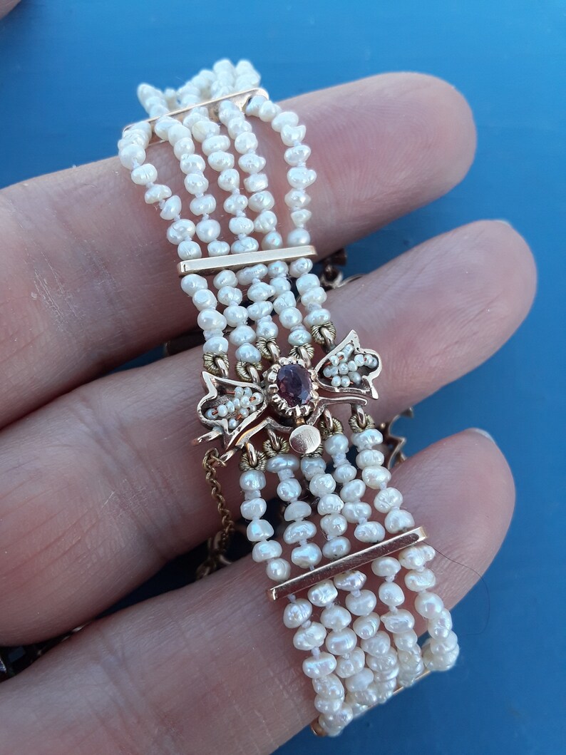K&ouml;nnte beinhalten: Ein goldenes Armband mit einem roten Edelstein und Perlenakzenten. Das Armband besteht aus mehreren Reihen von Perlen und hat einen dekorativen Verschluss.