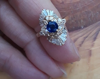 May include: A gold ring with a blue sapphire center stone surrounded by smaller diamonds. The ring has a delicate, intricate design.