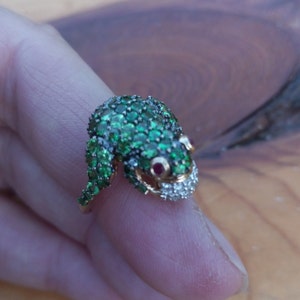 May include: A gold ring featuring a detailed frog design. The frog is set with green gemstones and has red ruby eyes. The frog's mouth is open and has a row of white diamonds.