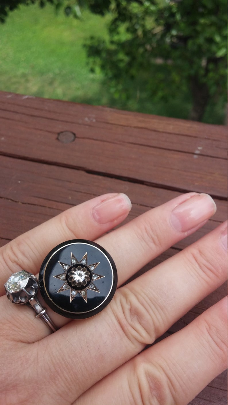 K&ouml;nnte beinhalten: Ein schwarzer Ring mit einer gro&szlig;en, runden, schwarzen Emaillefassung. Die Fassung zeigt ein Sternenmuster mit einem gro&szlig;en, klaren, runden Stein in der Mitte und kleineren, klaren Steinen am Rand.