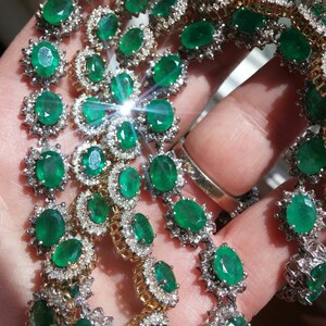 May include: A close-up of a multi-strand emerald and diamond necklace. The necklace features oval-shaped emeralds set in a halo of round diamonds. The emeralds are a vibrant green color and the diamonds are sparkling.