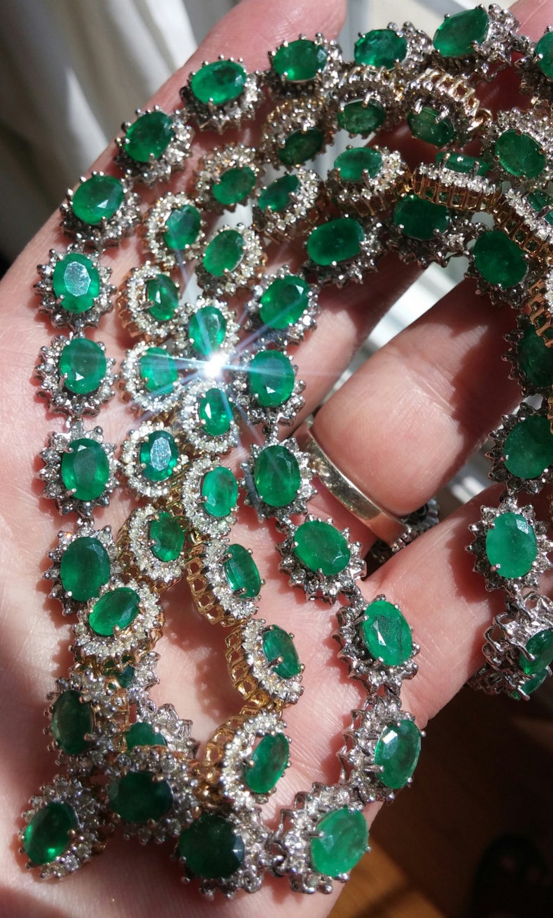 May include: A close-up of a multi-strand emerald and diamond necklace. The necklace features oval-shaped emeralds set in a halo of round diamonds. The emeralds are a vibrant green color and the diamonds are sparkling.