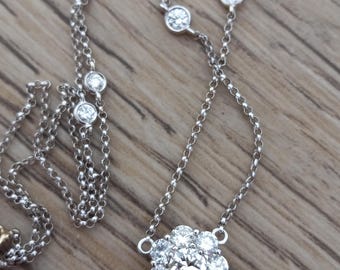 May include: A delicate silver necklace featuring a cluster of small, sparkling diamonds in a floral design. The necklace has a delicate chain with additional bezel-set diamonds. The jewelry is displayed on a wooden surface.