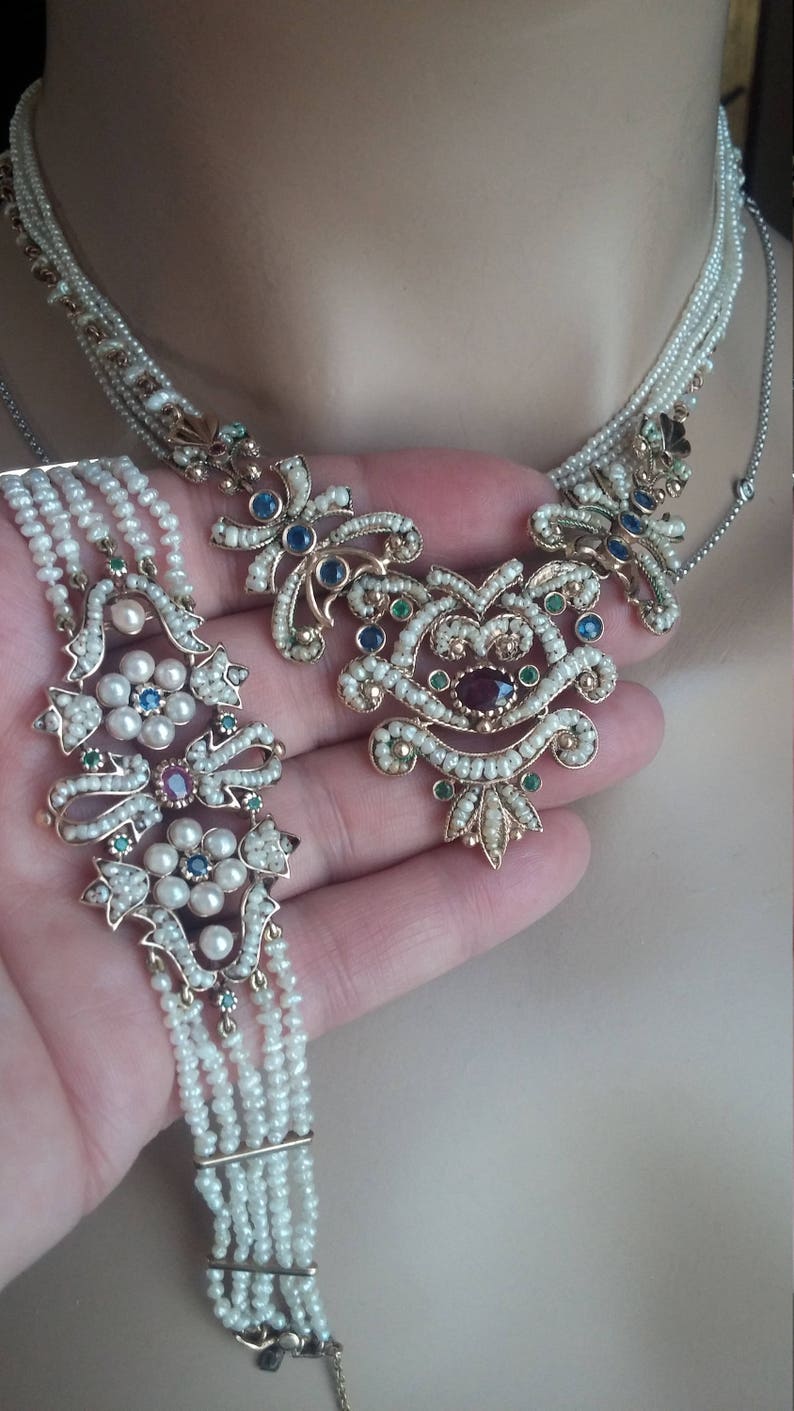 K&ouml;nnte beinhalten: Ein zartes Perlen-Halsketten- und Armband-Set mit filigranen Blumenmustern und bunten Edelsteinen. Die Halskette hat einen gro&szlig;en Anh&auml;nger mit einem Rubin in der Mitte, umgeben von Saphiren und Smaragden. Das Armband ist ein mehrreihiges Design mit einem &auml;hnlichen Blumenmotiv.