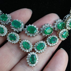 May include: A silver necklace with a large emerald pendant surrounded by smaller diamonds. The necklace is made up of a chain of smaller emeralds and diamonds.