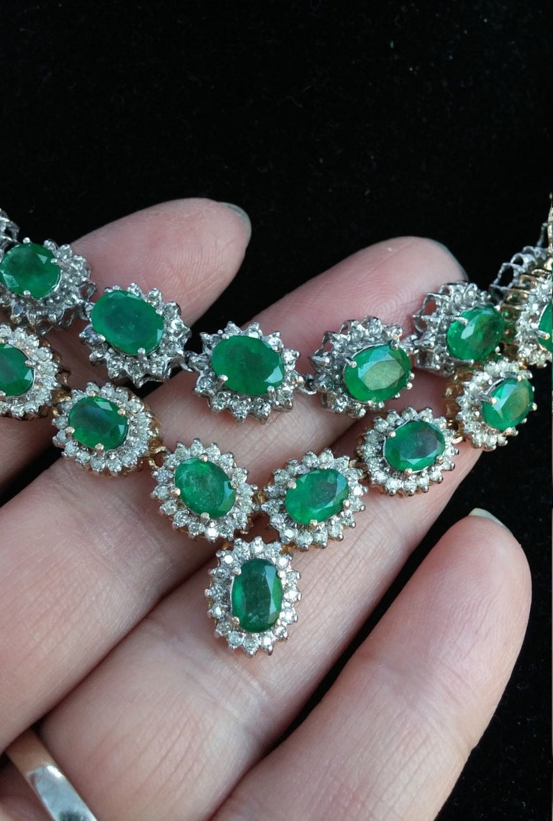 May include: A silver necklace with a large emerald pendant surrounded by smaller diamonds. The necklace is made up of a chain of smaller emeralds and diamonds.