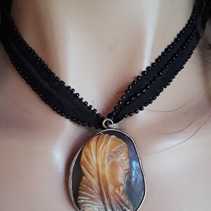 May include: A black beaded choker necklace with a cameo pendant featuring a carved profile of a woman wearing a head covering. The cameo is set in a silver-toned metal frame.