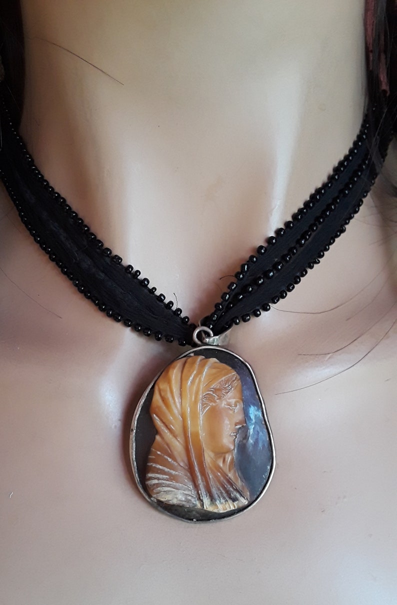 May include: A black beaded choker necklace with a cameo pendant featuring a carved profile of a woman wearing a head covering. The cameo is set in a silver-toned metal frame.