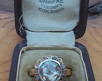 Puede incluir: Un anillo con una gran piedra preciosa facetada y transparente rodeada de piedras preciosas más pequeñas y transparentes engastadas en una banda de oro. El anillo está en una caja de terciopelo marrón con la inscripción "FABERGE  C.C. ПЕТЕРБУРГЪ  МОСКВА, ЛОНДОНЪ".