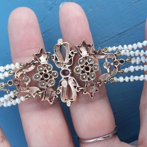 K&ouml;nnte beinhalten: Ein goldenes Armband mit einem gro&szlig;en, verzierten Mittelst&uuml;ck, das mit roten und wei&szlig;en Edelsteinen besetzt ist. Das Armband besteht aus mehreren Reihen Perlen.