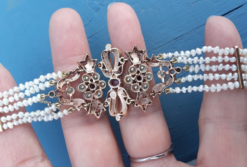 K&ouml;nnte beinhalten: Ein goldenes Armband mit einem gro&szlig;en, verzierten Mittelst&uuml;ck, das mit roten und wei&szlig;en Edelsteinen besetzt ist. Das Armband besteht aus mehreren Reihen Perlen.