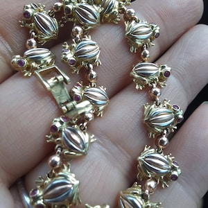 May include: A gold-toned bracelet featuring a repeating frog design. Each frog has a ribbed body with a copper-colored center and small red gemstone eyes. The bracelet is secured with a clasp.