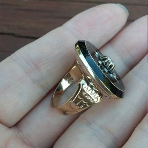 K&ouml;nnte beinhalten: Ein goldener Ring mit einem schwarzen ovalen Cabochon in der Mitte. Der Cabochon ist von kleinen Diamanten umgeben. Die Ringband ist mit einem floralen Muster verziert.