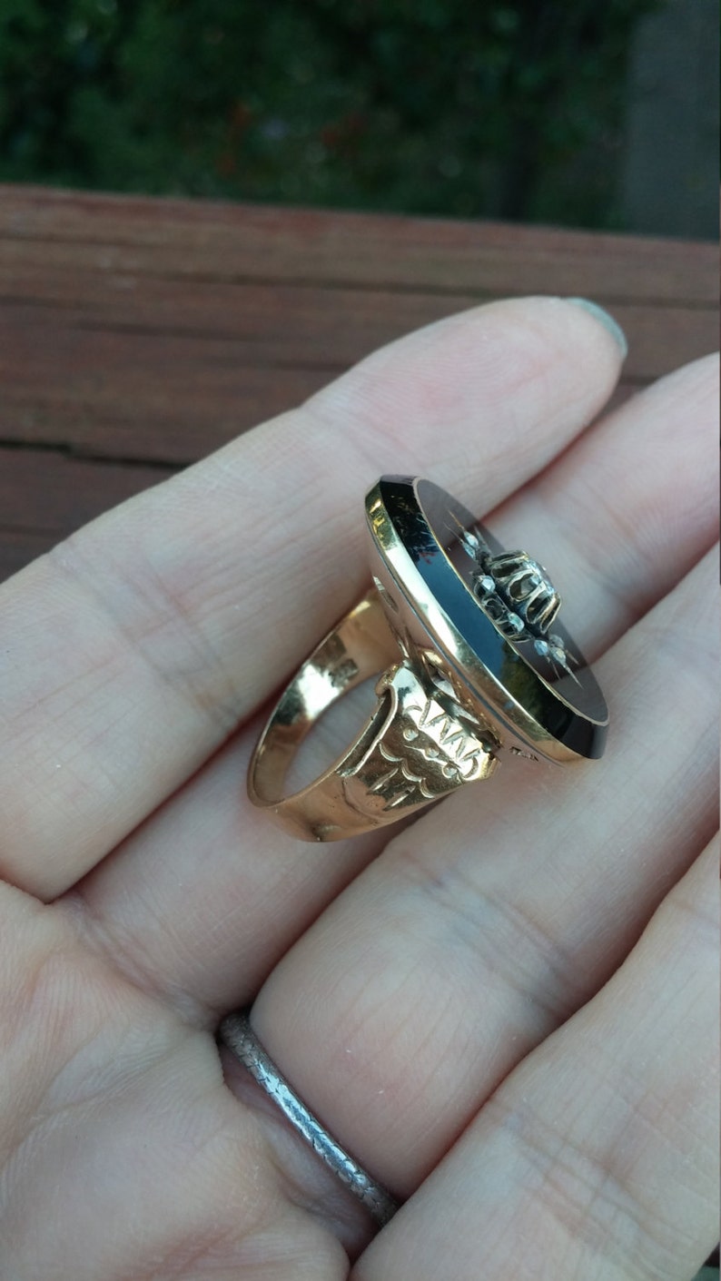 K&ouml;nnte beinhalten: Ein goldener Ring mit einem schwarzen ovalen Cabochon in der Mitte. Der Cabochon ist von kleinen Diamanten umgeben. Die Ringband ist mit einem floralen Muster verziert.