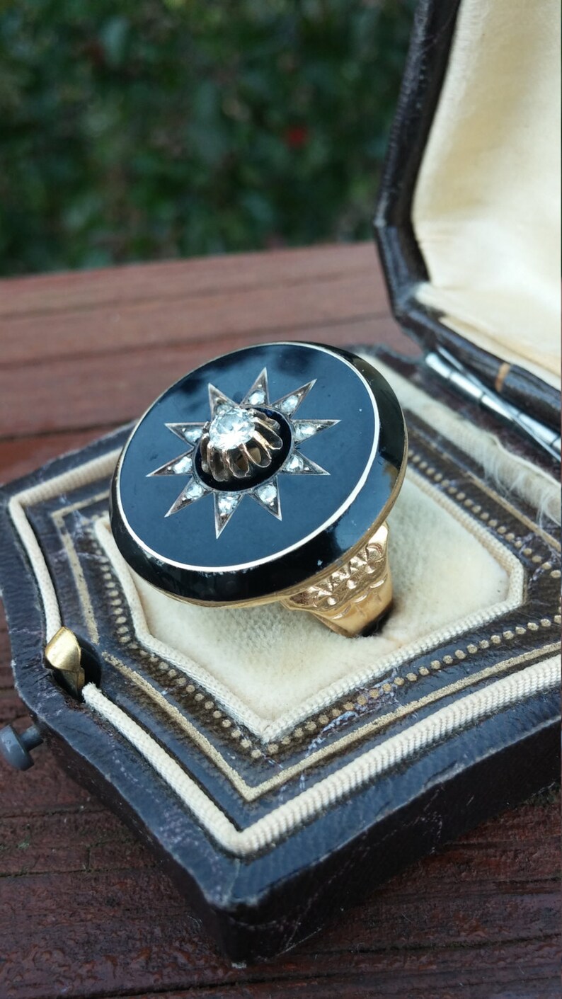 K&ouml;nnte beinhalten: Ein schwarzer Emailring mit einem Goldband und einem gro&szlig;en, klaren Diamanten, der in einem Sternendesign gefasst ist.