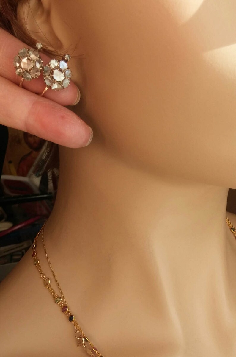 May include: A pair of gold earrings with a cluster of small, clear stones. The earrings are on a mannequin's ear. The mannequin is also wearing a gold necklace with small, colourful stones.