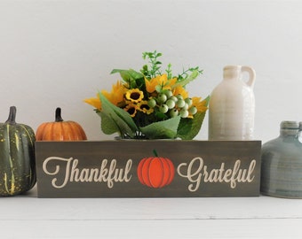 May include: A wooden sign with the words "Thankful" and "Grateful" in a cursive font. A small orange pumpkin is between the words. The sign is sitting on a white surface with a small bouquet of sunflowers and a ceramic pitcher.