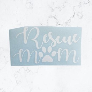 May include: A light blue sticker with the words "Rescue Mom" in white cursive script. A white paw print graphic is between the words "Rescue" and "Mom". The sticker is rectangular and sits on a white marble surface.