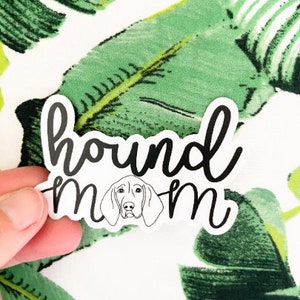 May include: A white sticker with the words "hound mom" in black script, featuring a line drawing of a dog's face. The sticker is held against a background of green and white tropical leaves.