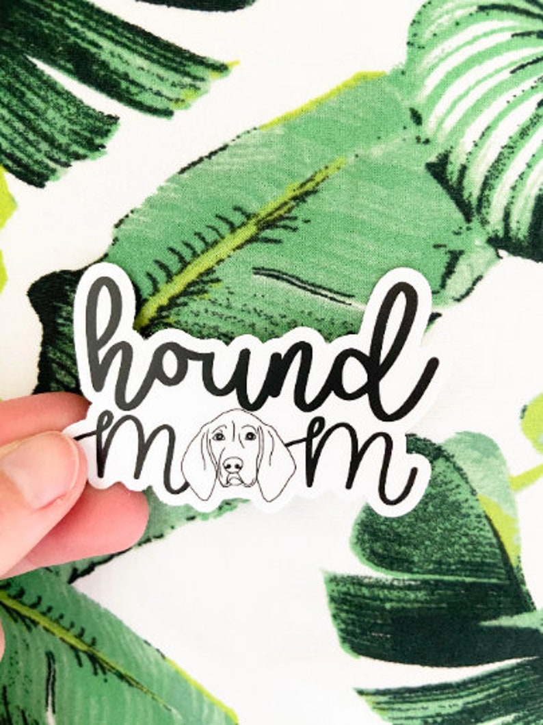 May include: A white sticker with the words "hound mom" in black script, featuring a line drawing of a dog's face. The sticker is held against a background of green and white tropical leaves.