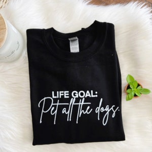 K&ouml;nnte beinhalten: Schwarzes Sweatshirt mit dem wei&szlig;en Text "LIFE GOAL: Pet all the dogs". Das Sweatshirt ist auf einer wei&szlig;en, flauschigen Oberfl&auml;che gefaltet, mit einer Kerze und einer kleinen Topfpflanze im Hintergrund.