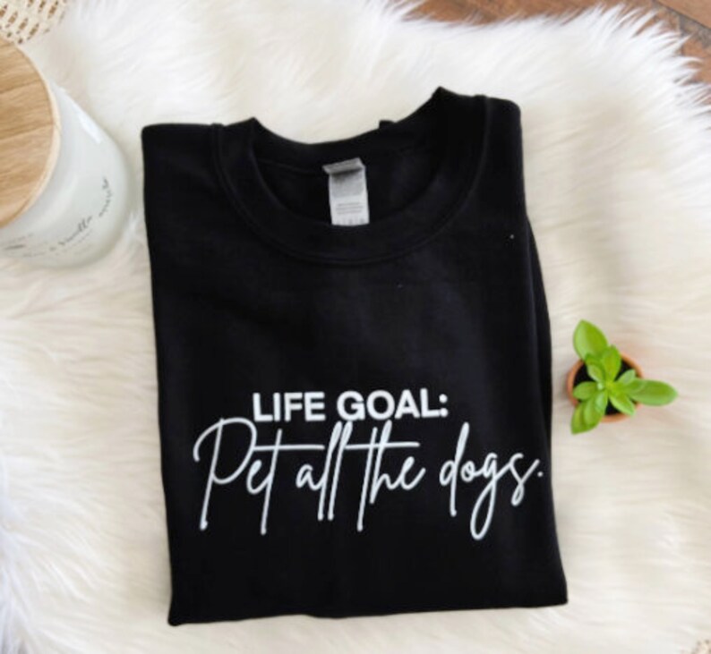 K&ouml;nnte beinhalten: Schwarzes Sweatshirt mit dem wei&szlig;en Text "LIFE GOAL: Pet all the dogs". Das Sweatshirt ist auf einer wei&szlig;en, flauschigen Oberfl&auml;che gefaltet, mit einer Kerze und einer kleinen Topfpflanze im Hintergrund.
