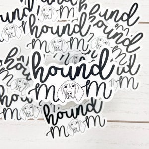 May include: A collection of white stickers with a black script font that reads "hound mom." Each sticker features a line drawing of a hound dog's face. The stickers are scattered on a white wooden surface.