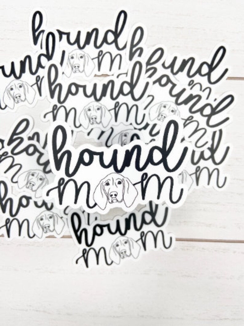 May include: A collection of white stickers with a black script font that reads "hound mom." Each sticker features a line drawing of a hound dog's face. The stickers are scattered on a white wooden surface.