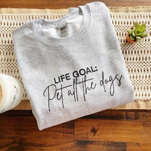 K&ouml;nnte beinhalten: Ein meliertes graues Sweatshirt mit dem Text "LIFE GOAL: Pet all the dogs" in schwarzer Schrift. Das Sweatshirt liegt gefaltet auf einem beigefarbenen, gewebten Tischl&auml;ufer auf einer Holzoberfl&auml;che. Eine kleine Topf-Sukkulente ist sichtbar.