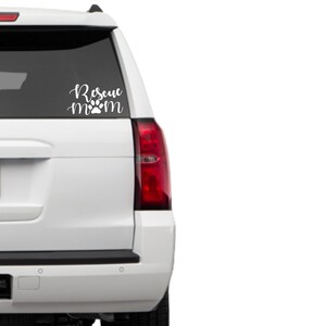 May include: White car window decal with the words "Rescue Mom" in a cursive font, with a paw print replacing the "o" in "mom". The decal is designed to be applied to a vehicle's rear window.